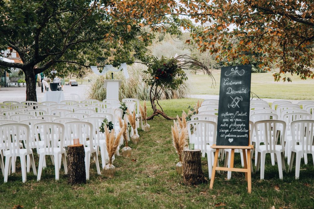 Outdoor wedding ceremony setup with white chairs, wooden stumps, and a welcome chalkboard sign on an easel.