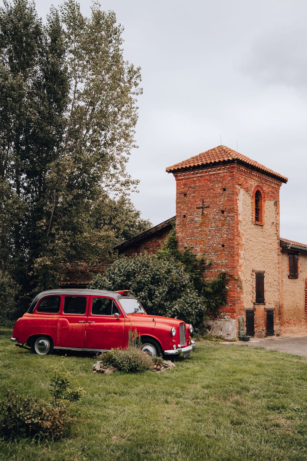 Vintage red classic car parked beside a rustic brick chapel tower with cross in a green countryside setting.