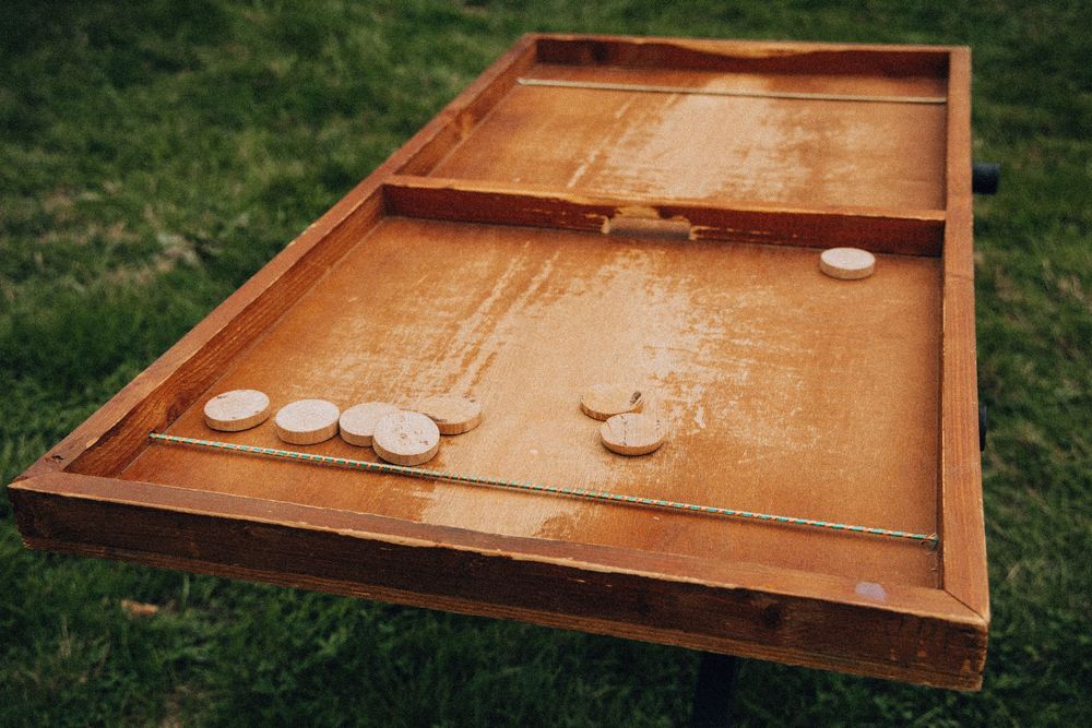 Rustic wooden shuffleboard game table with white pucks on green grass at an outdoor wedding reception.