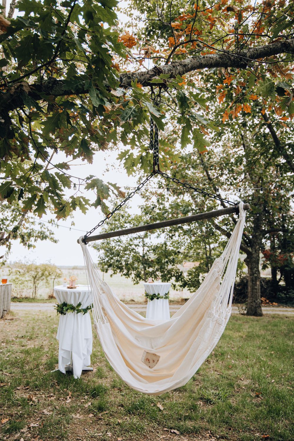 Bohemian macrame hammock swing hanging from tree branch with white draped reception tables in garden background.