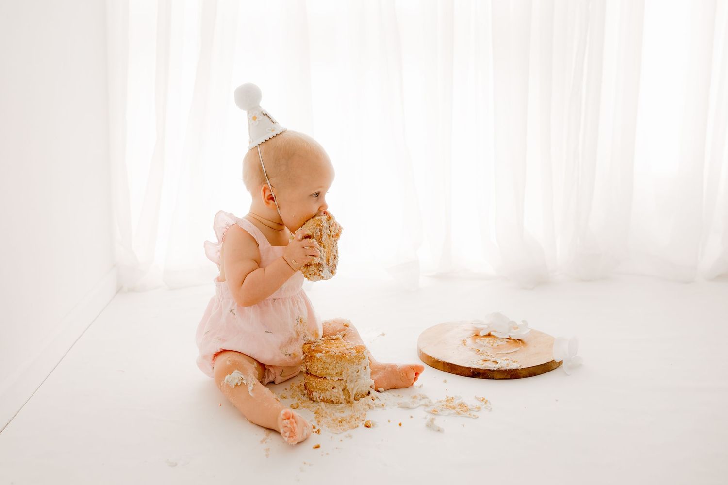 Baby in white outfit playing with cake and frosting during cake smash photo session.