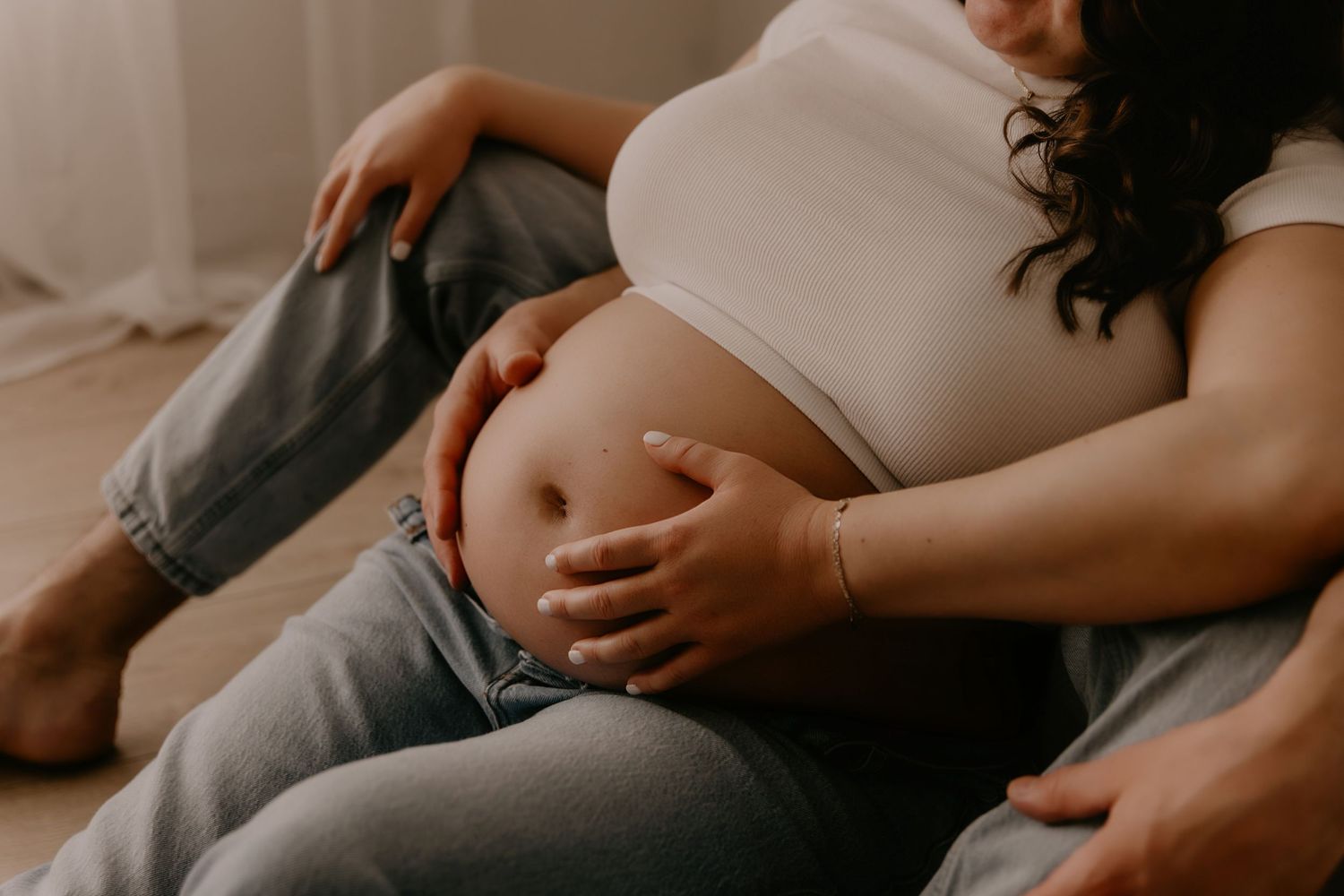 Pregnant woman in white crop top and jeans cradling her bare baby bump while sitting on the floor.