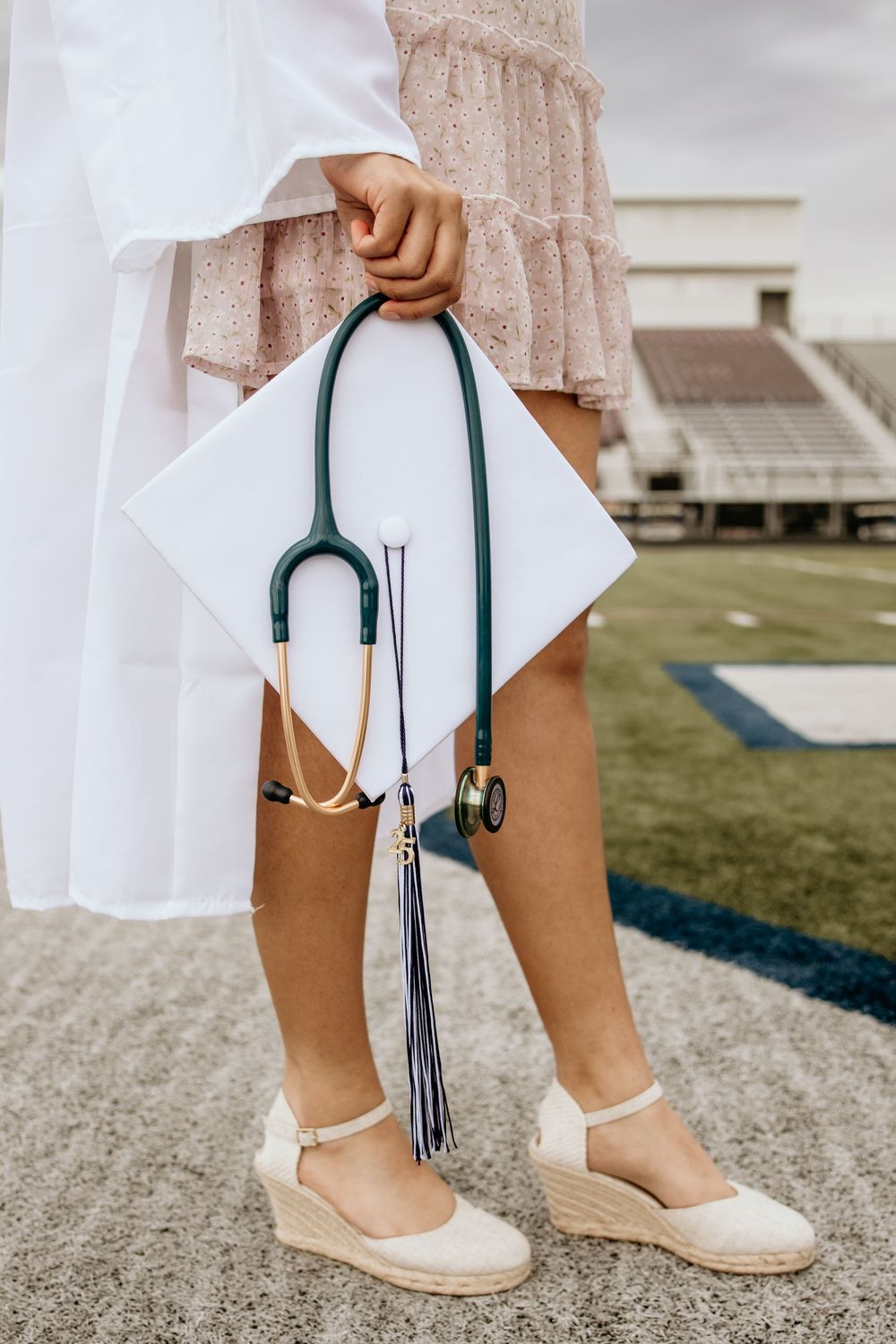 A nursing graduate holds a white cap and teal stethoscope while wearing espadrille wedges at a stadium.
