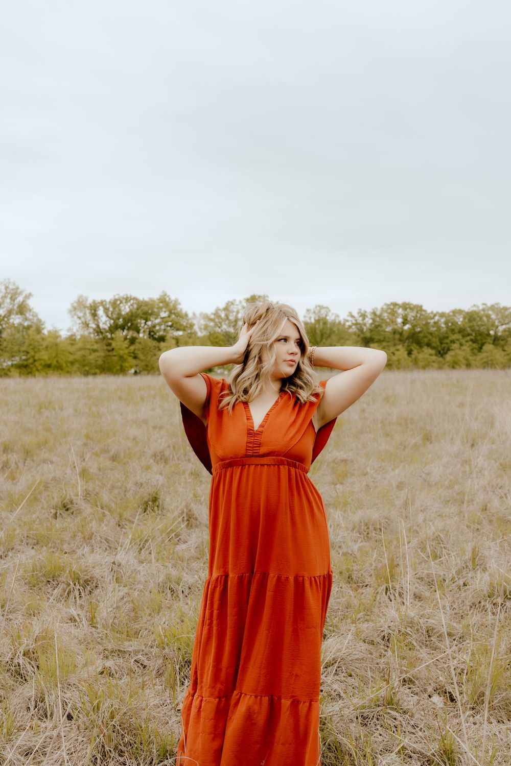 Young blonde woman in a flowing rust-orange maxi dress stands in an open field with hands in hair.