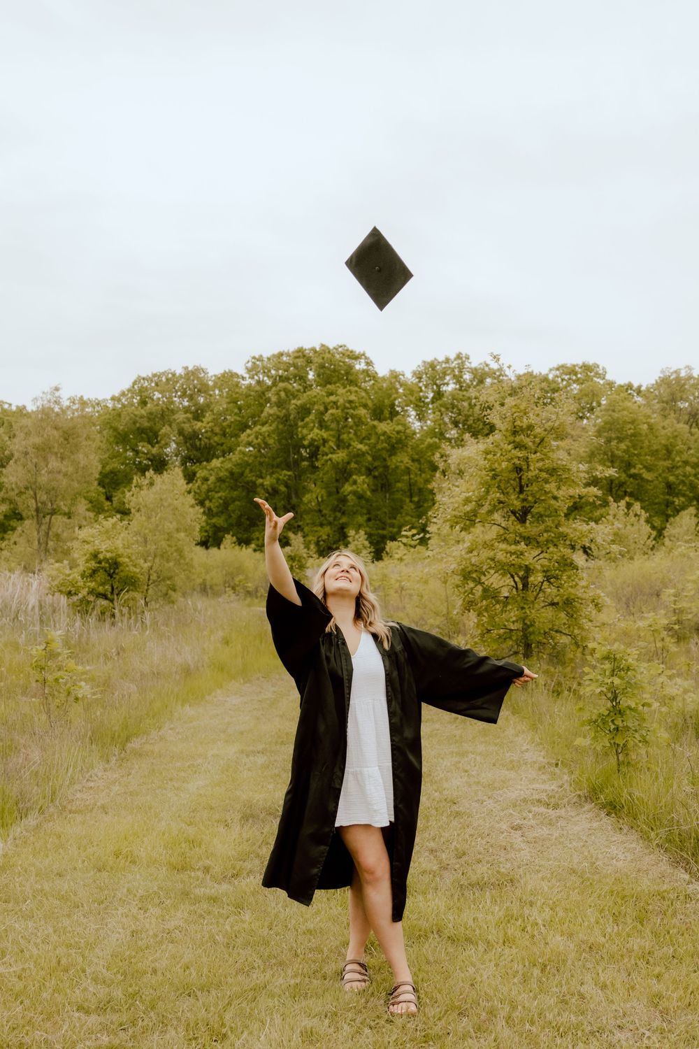 A smiling graduate in black gown throws her mortarboard cap into the air outdoors.
