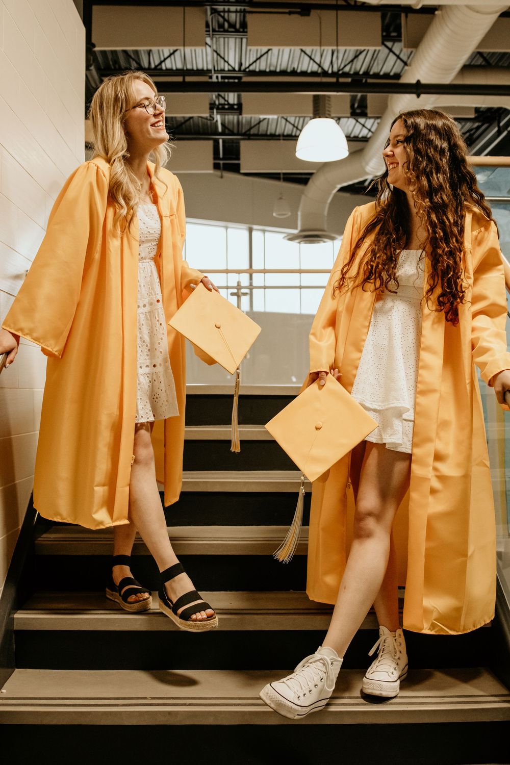Two female graduates in gold caps and gowns laughing on stairs during graduation day.