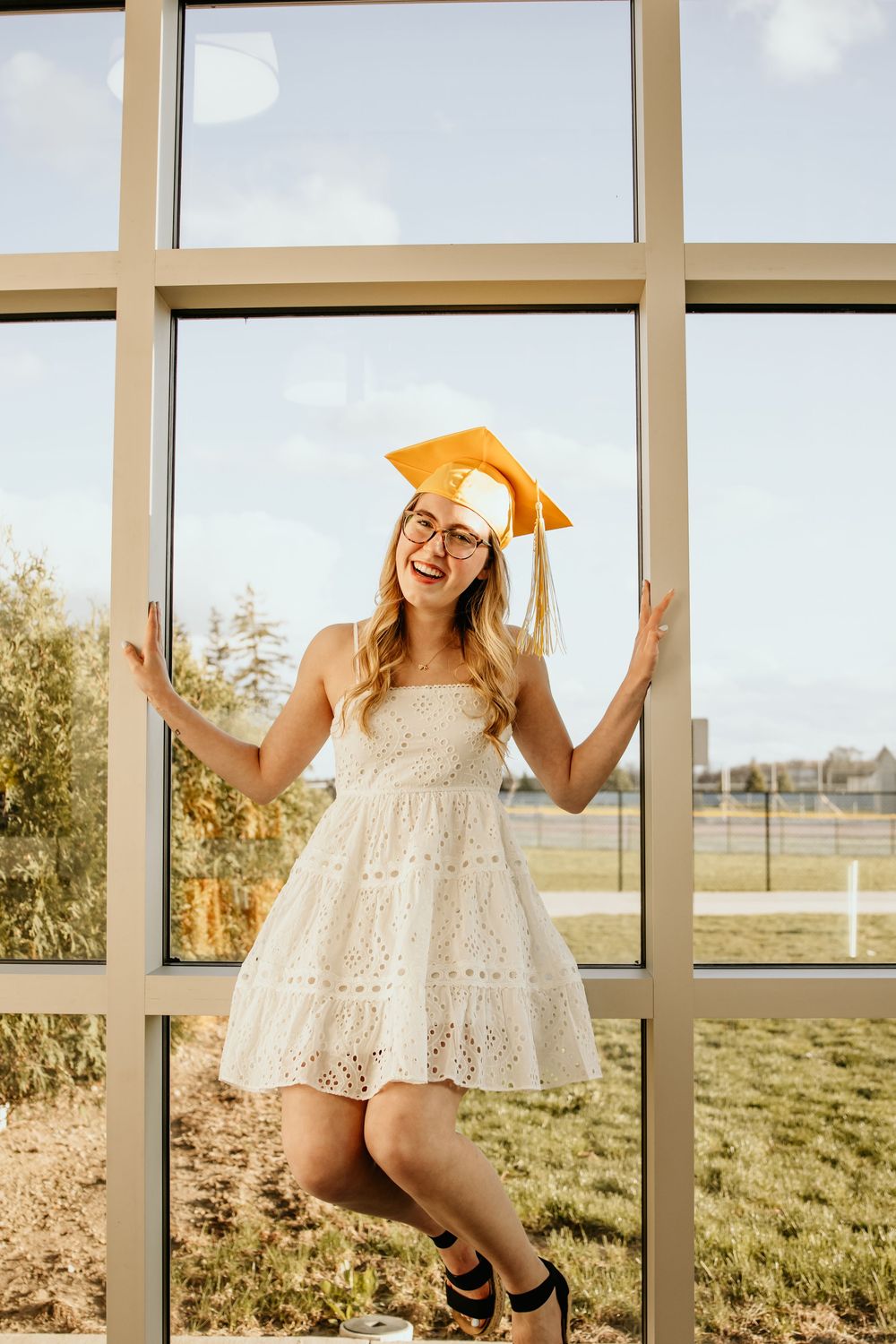 Happy female graduate in white dress and gold mortarboard posing by large school windows.