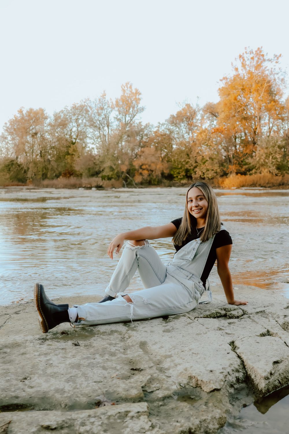 Smiling teenage girl in white overalls sits on rocks by river for fall senior portraits.