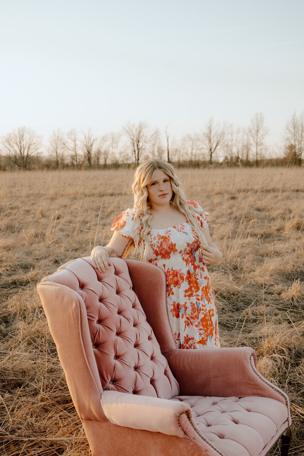 Pregnant woman in floral maternity dress standing behind a pink tufted chair in a golden field.