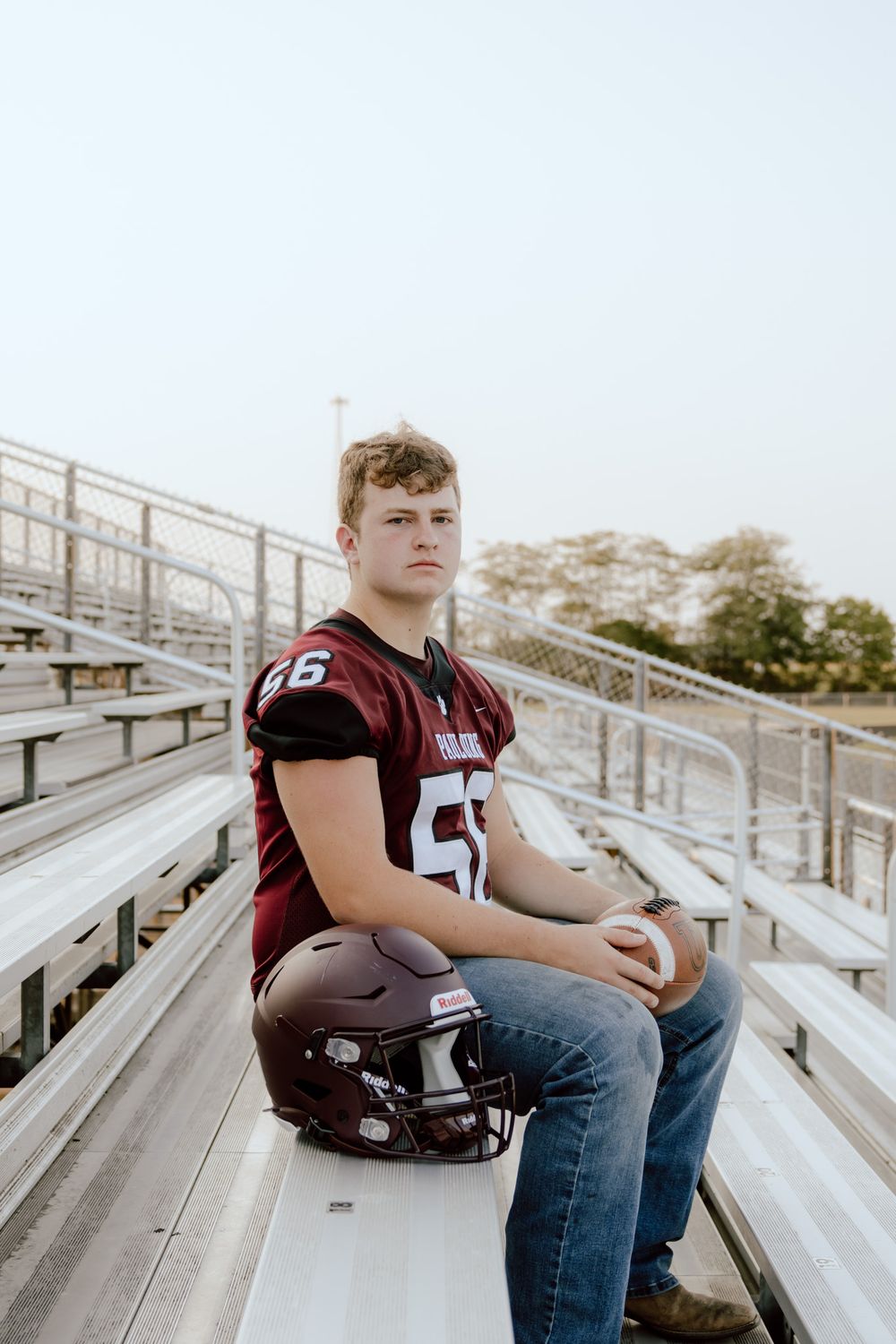 High school football player in Paulding #56 jersey sits in bleachers holding helmet and football.