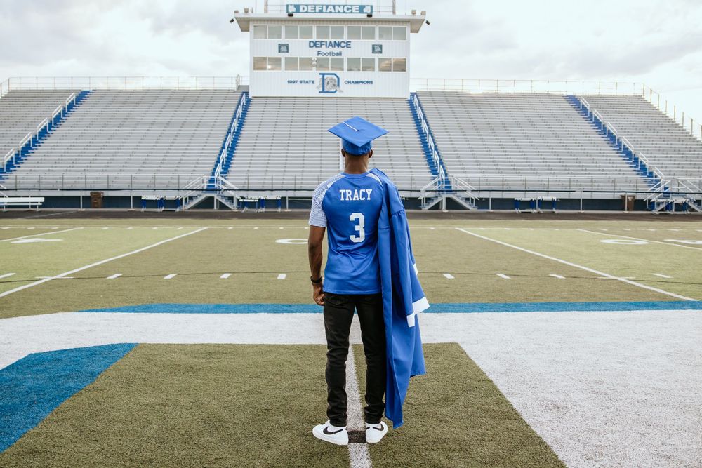 Graduate in blue cap and gown standing on football field at Defiance stadium.