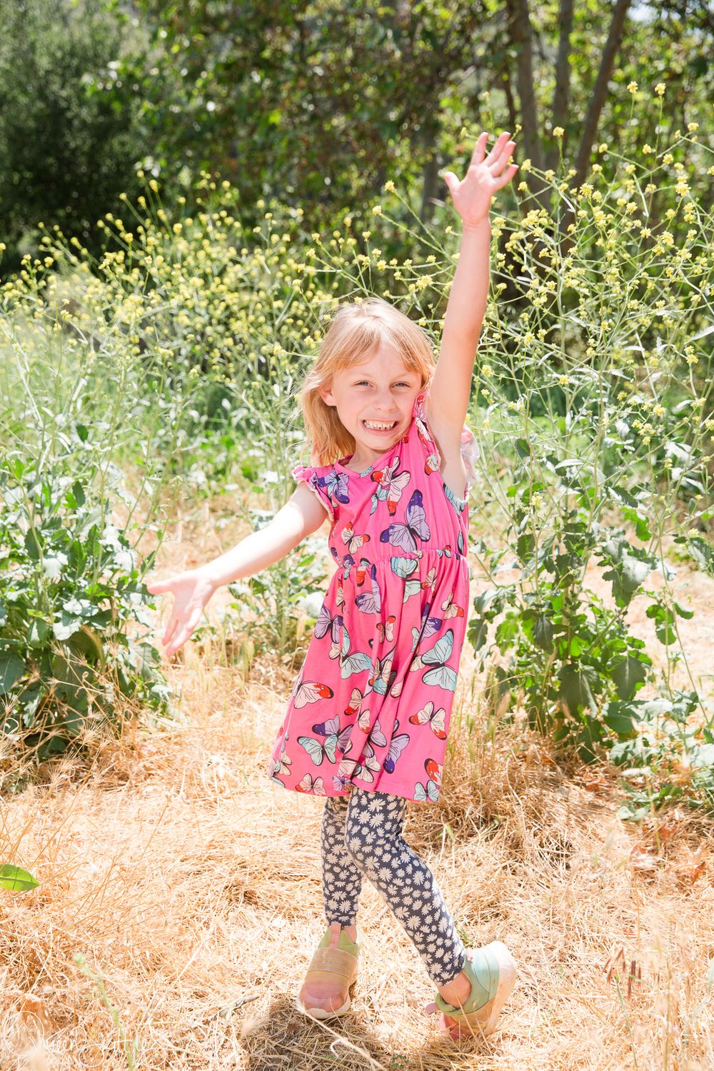 A child wearing a pink floral dress and patterned leggings dances joyfully in a sunny garden setting.