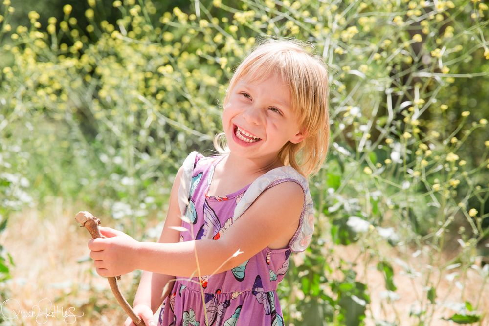 A young child in a purple dress plays joyfully outdoors among white wildflowers on a sunny summer day.