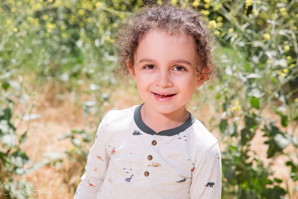 A child in a beige sweater smiles while standing outdoors near green foliage.