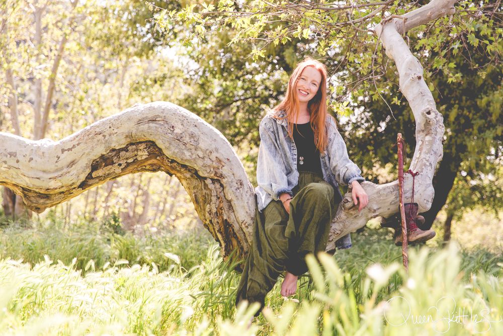 A couple sits together in a sun-dappled meadow surrounded by tall grass and trees during a romantic outdoor photo session.