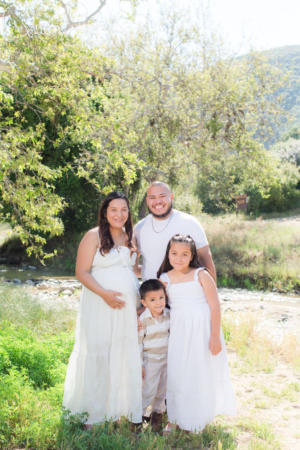 A family poses together outdoors in white clothing against a natural backdrop with trees and greenery.