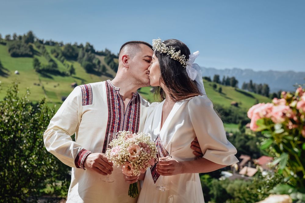 A couple in traditional white folk costumes embraces outdoors against a scenic mountain landscape on a sunny day.