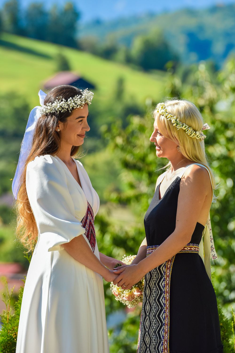 Two people in white and black dresses share joyful moments against a scenic mountain landscape with green hills.
