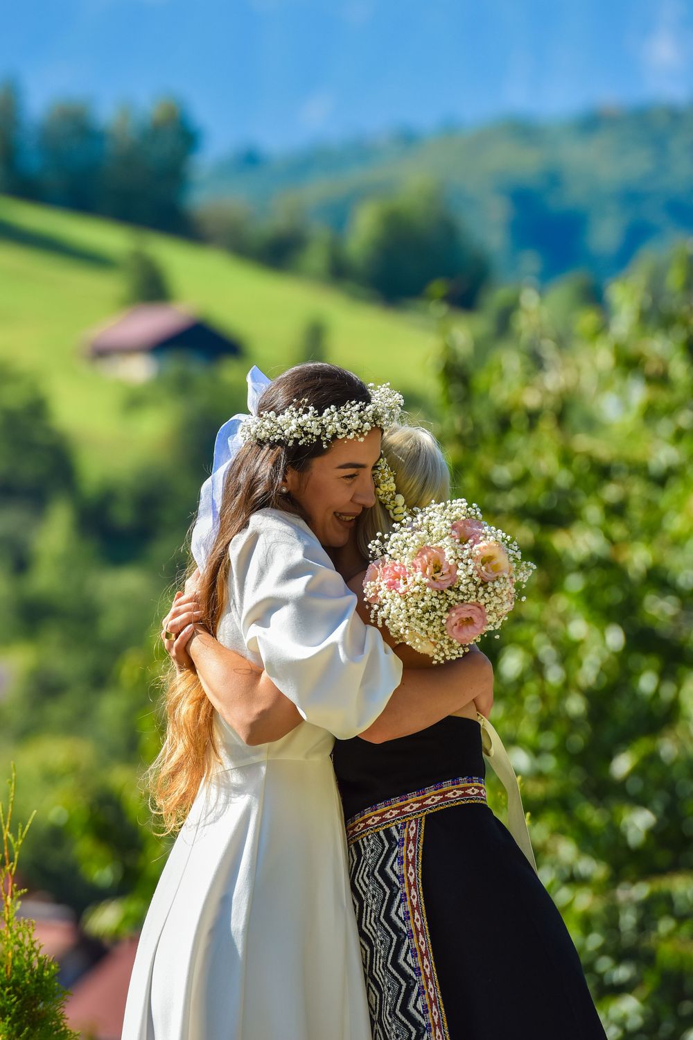 Two people embrace in a scenic vineyard setting with rolling green hills and mountains in the background.