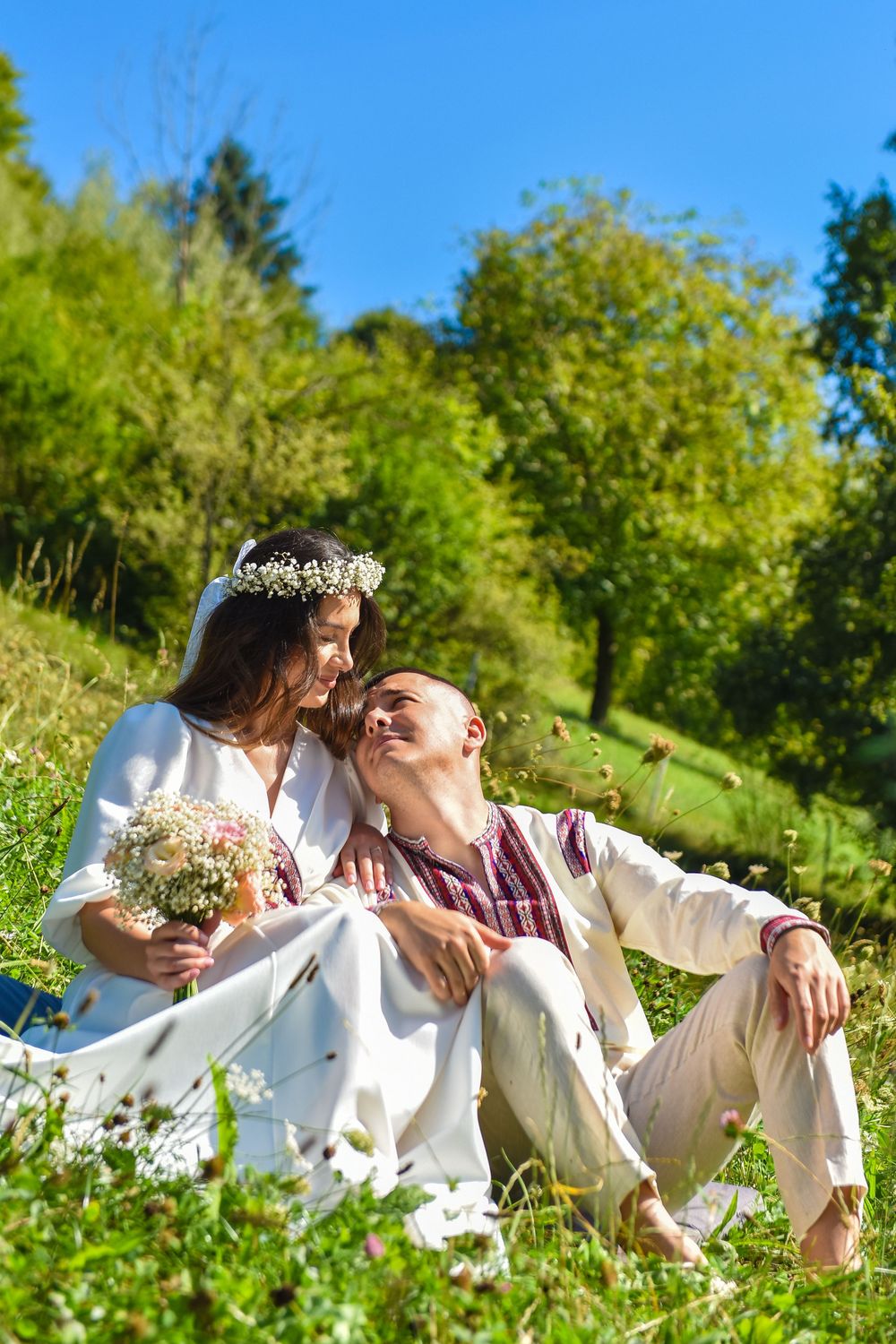 A couple in white clothing lies together in a sunny meadow surrounded by wildflowers and green grass.
