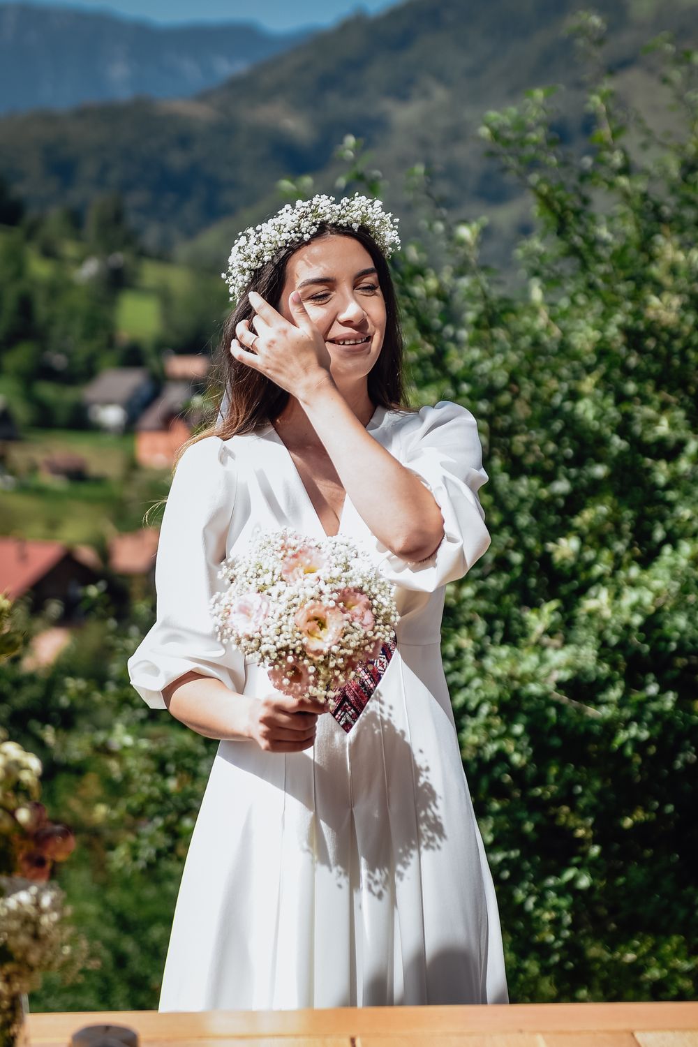 A person in a flowing white dress holds a delicate bouquet of flowers against a scenic mountain backdrop.