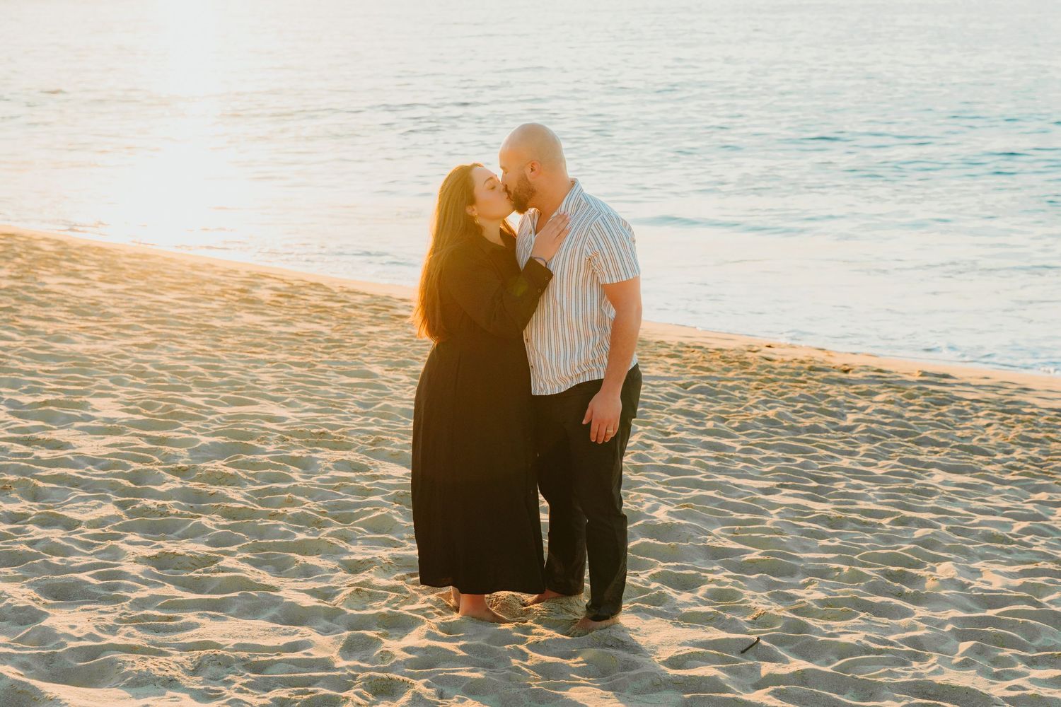A couple shares an intimate moment standing in the ocean waves at sunset with mountain peaks visible in the distance.