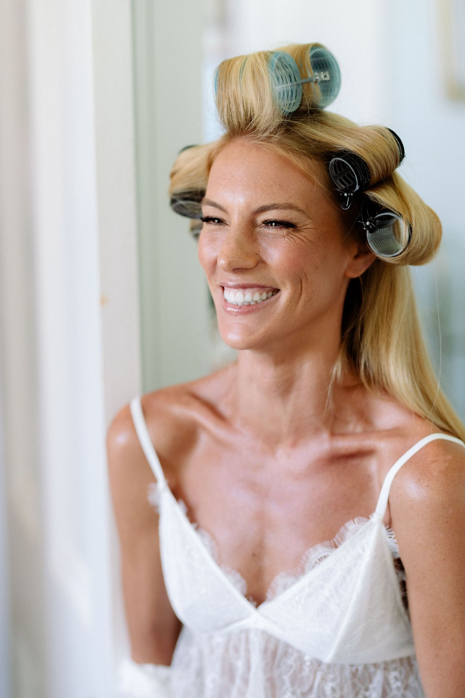 Bride getting ready with hair in rollers while wearing white lingerie and smiling.