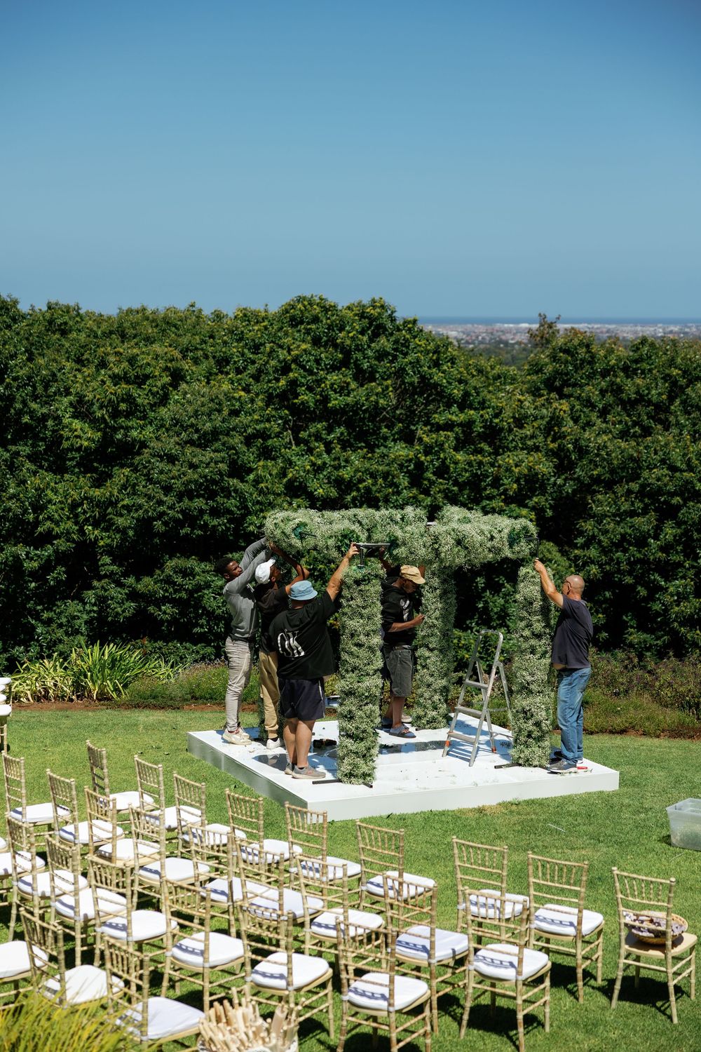 Workers set up a floral wedding arch on a platform overlooking green trees with gold chairs arranged for an outdoor ceremony.