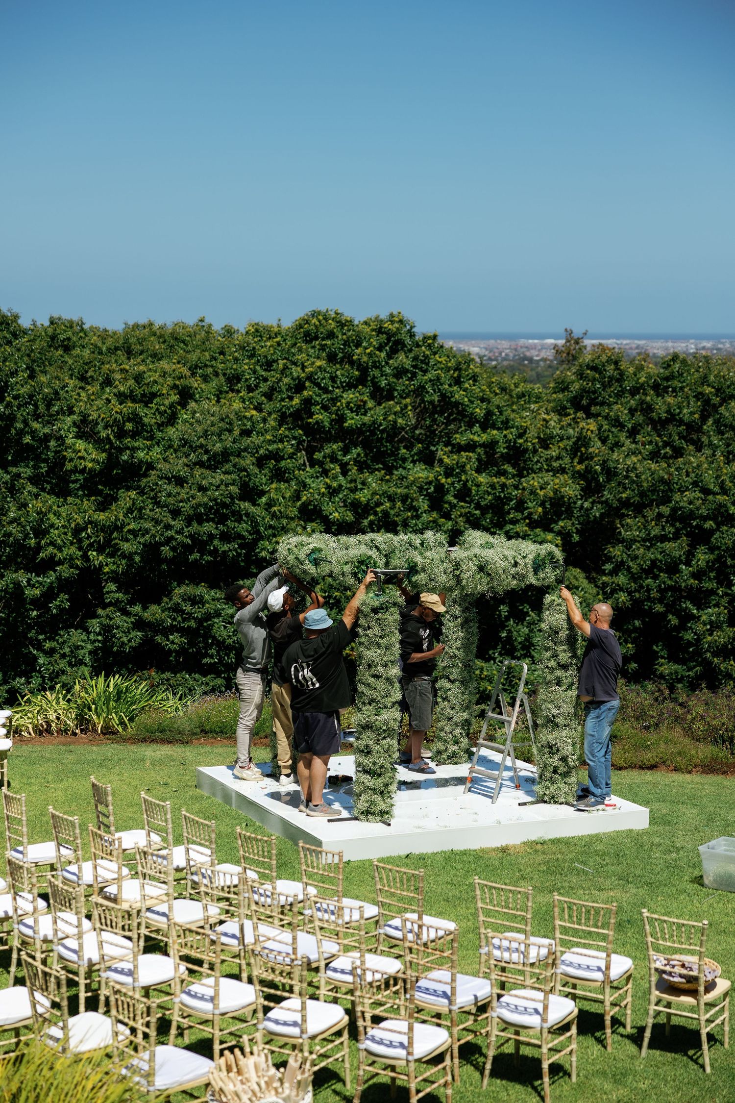 Workers set up a floral wedding arch on a platform overlooking green trees with gold chairs arranged for an outdoor ceremony.