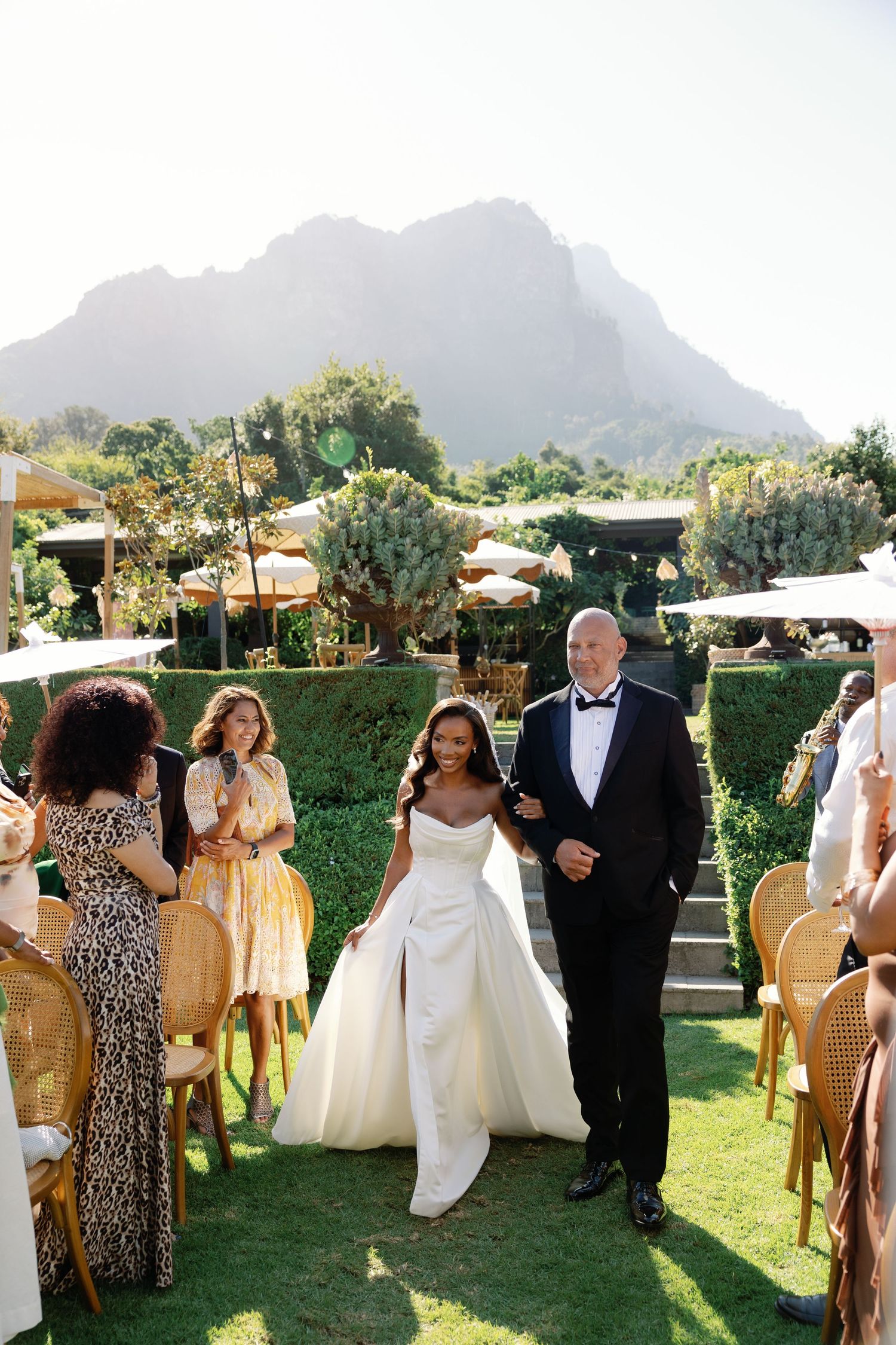 A wedding ceremony processional takes place on a lush lawn with majestic mountains in the background.