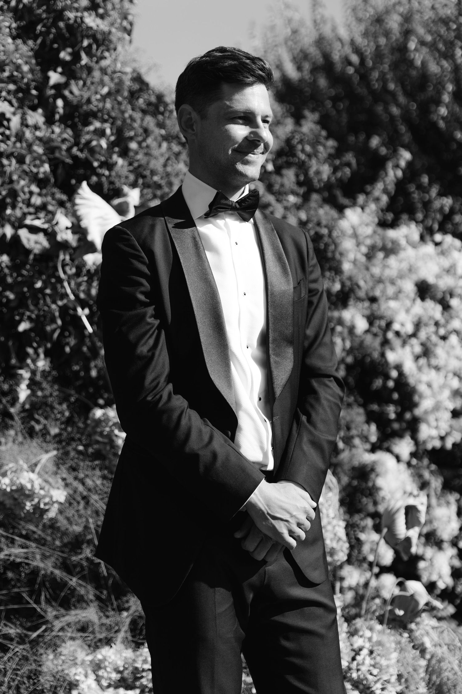 Formal wedding party in black tuxedos poses against a natural outdoor backdrop.