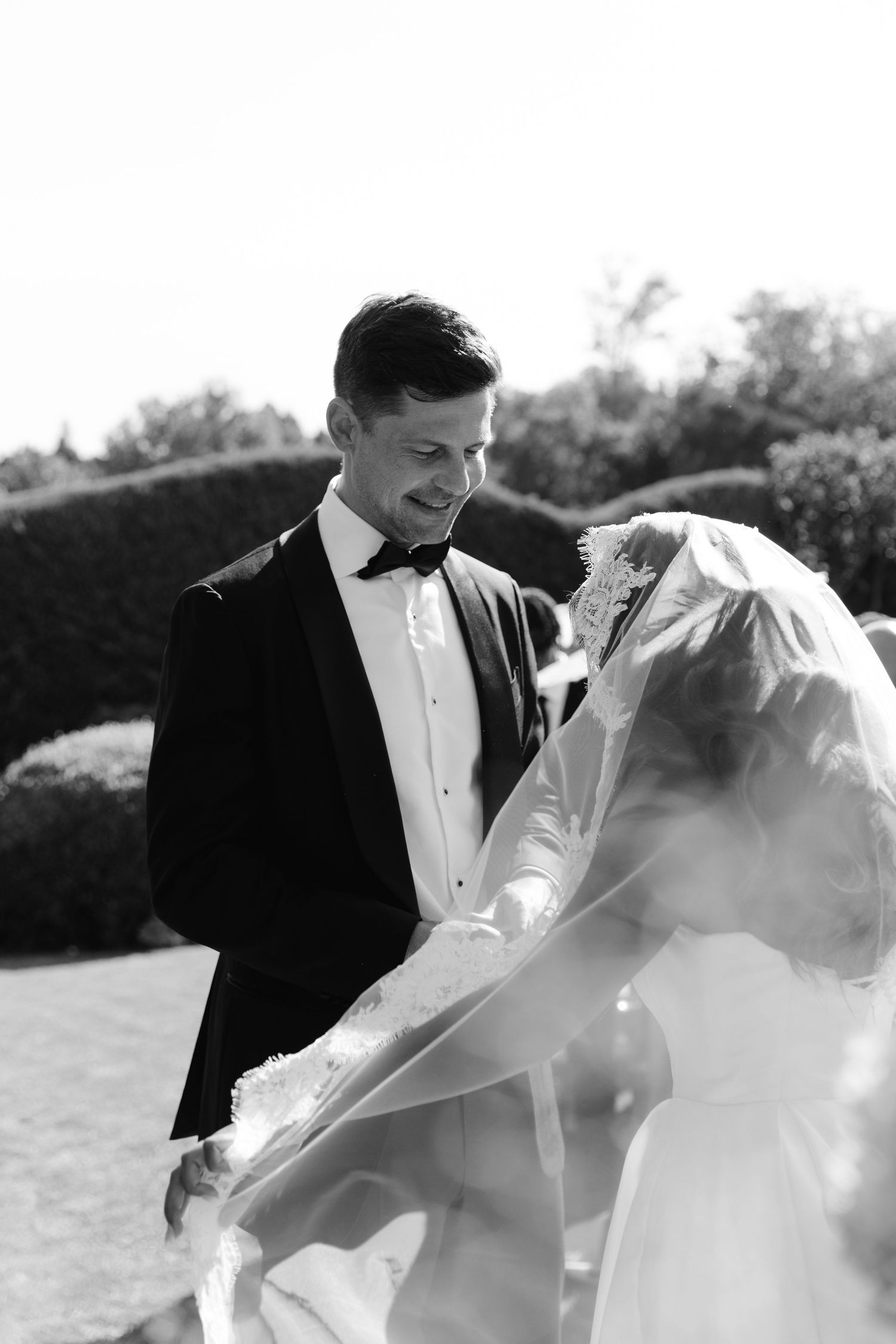 A romantic wedding couple shares an intimate moment under a flowing veil in black and white.