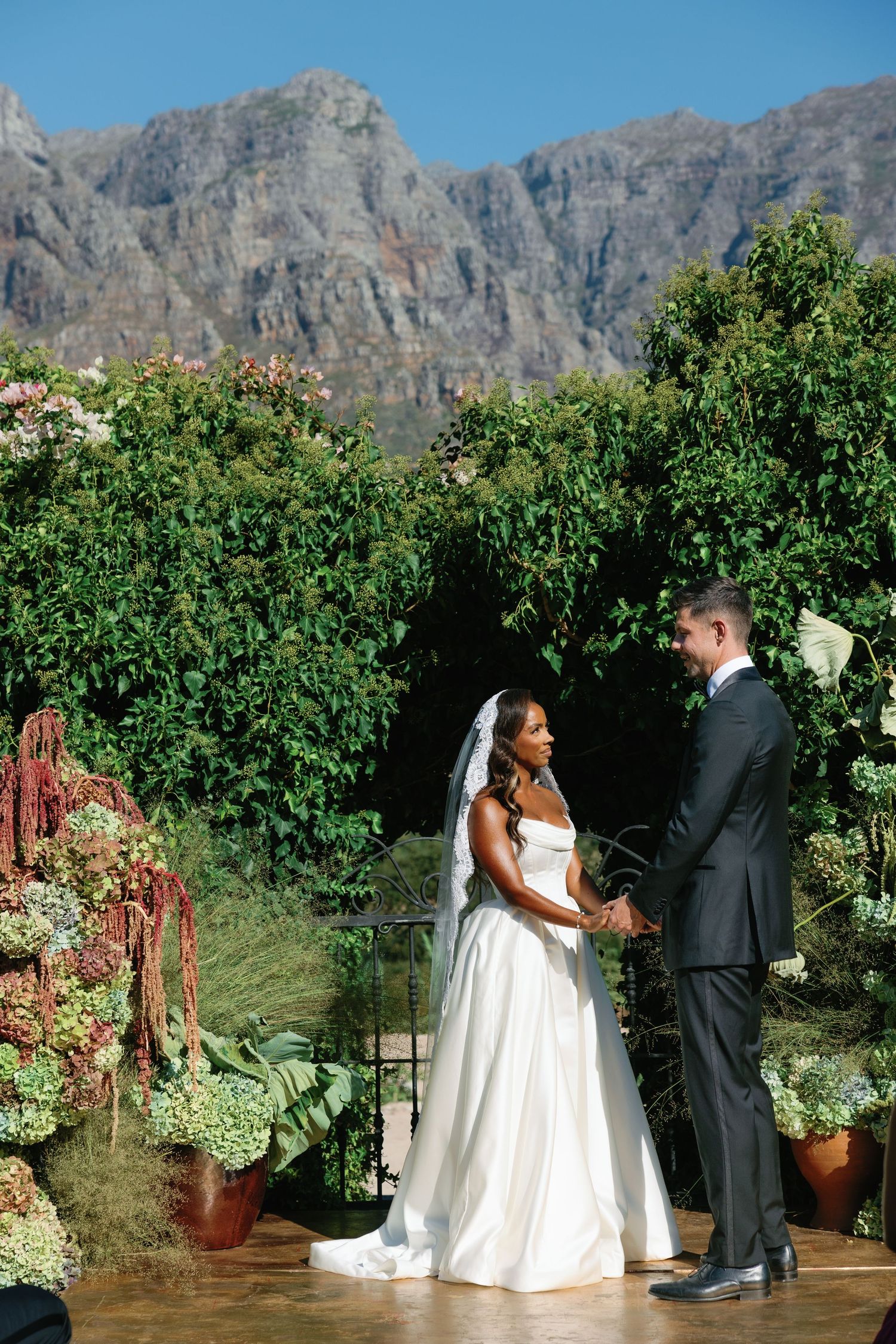 A wedding ceremony takes place in a garden with dramatic mountain peaks as the backdrop.