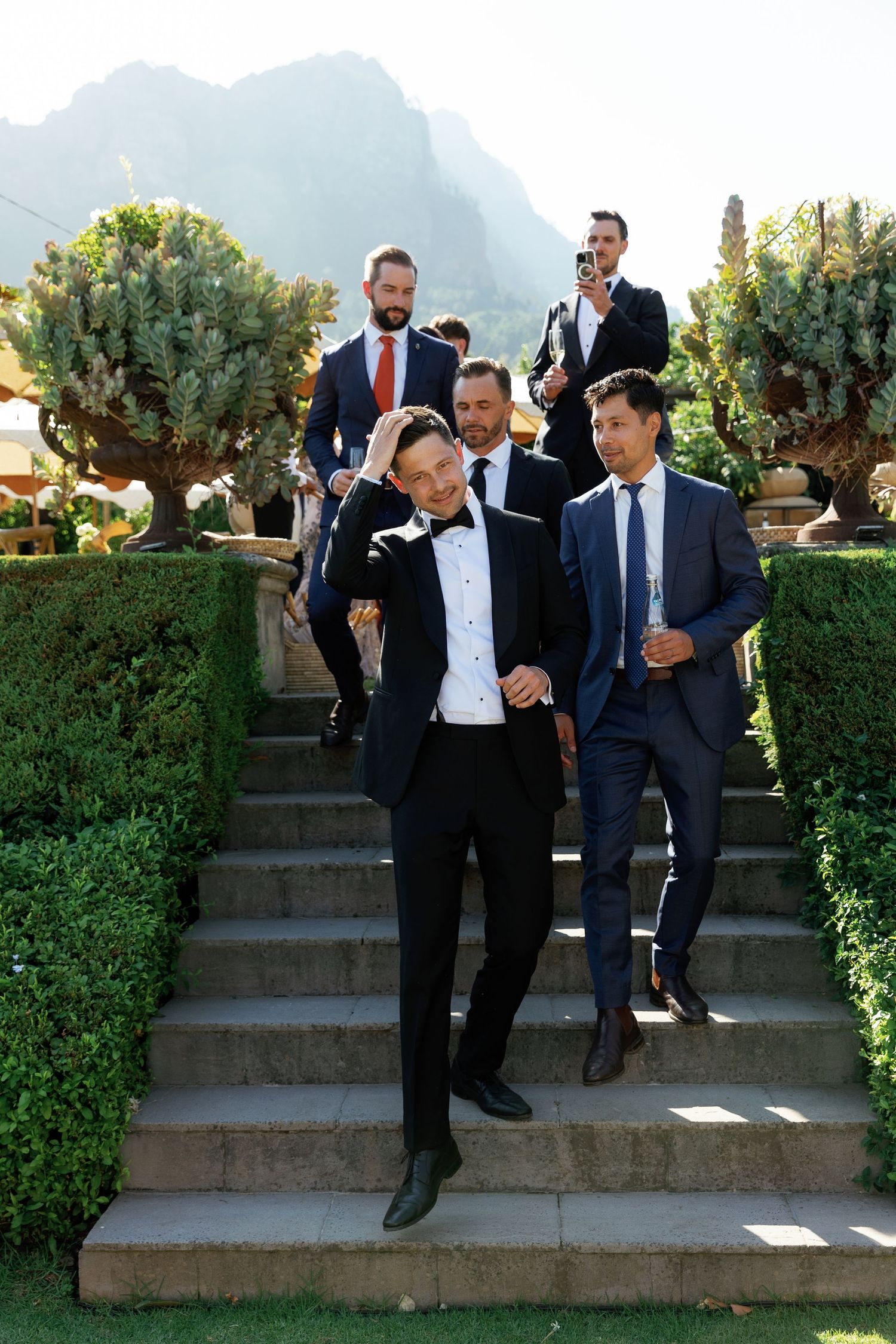 Groomsmen in dark suits pose playfully on outdoor stone steps surrounded by greenery.