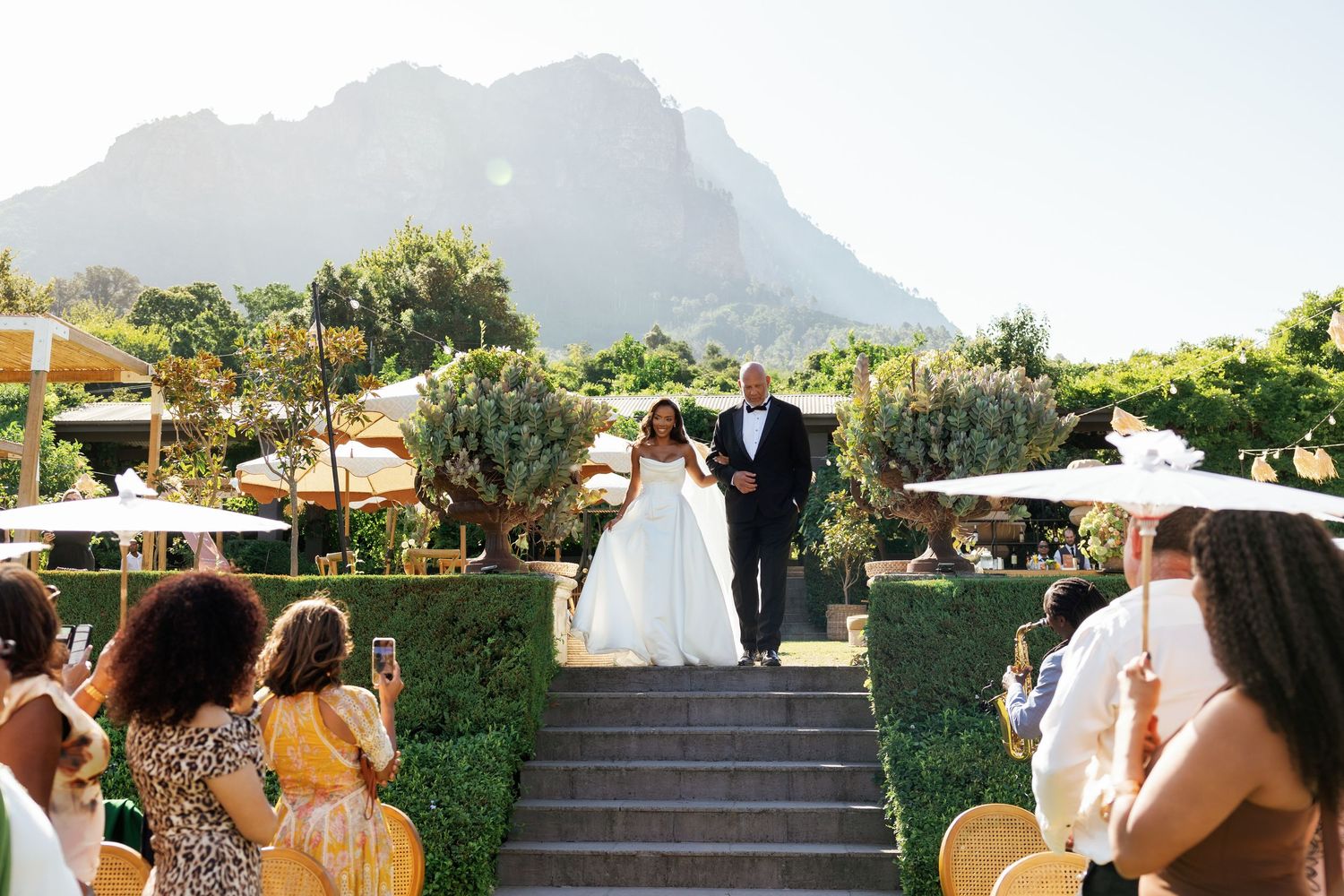 Outdoor wedding ceremony with guests seated on white chairs facing a mountain backdrop in a lush garden setting.