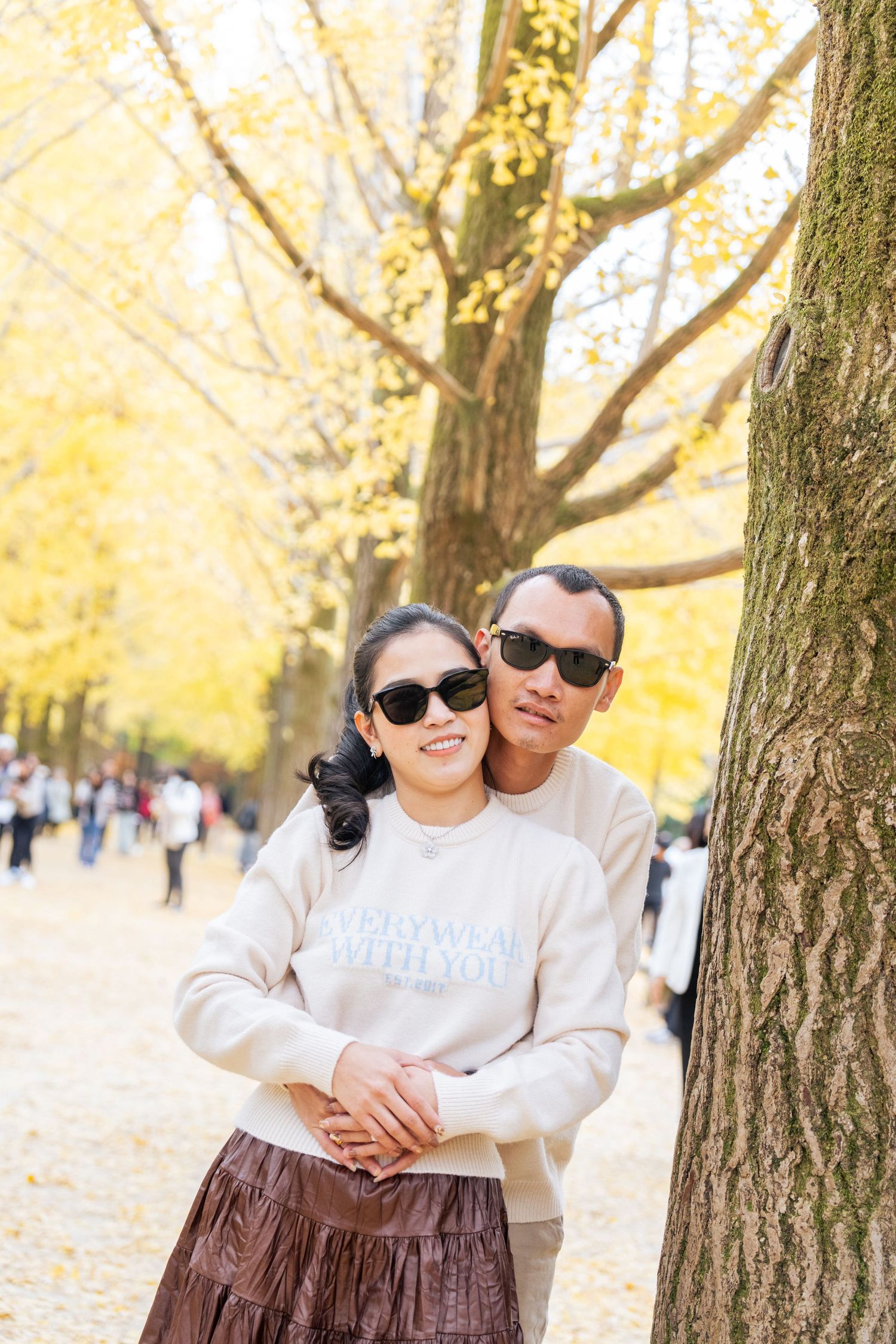 A couple embraces outdoors in a park during autumn with golden leaves and trees in the background.
