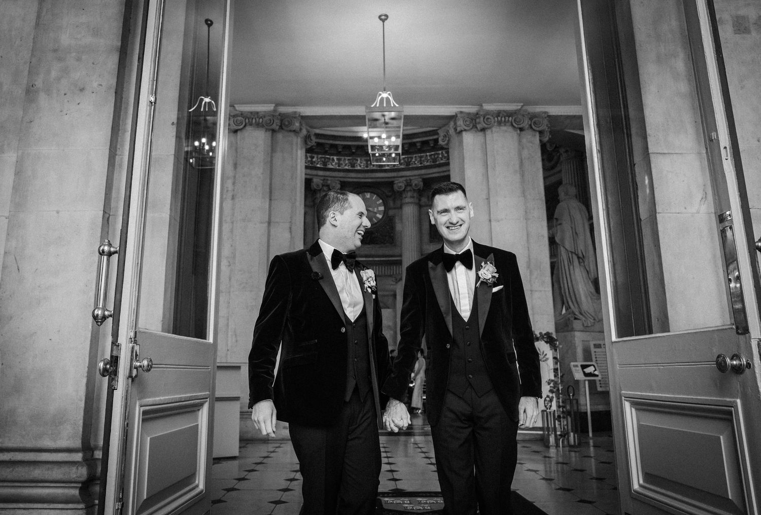 Two people in formal black attire stand together in an elegant hallway with white walls and ornate molding.
