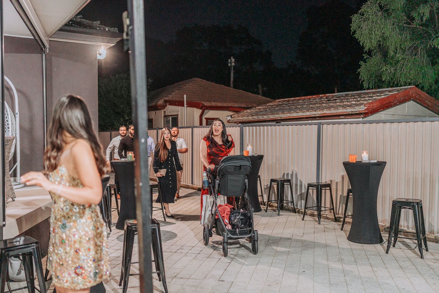 Evening outdoor party scene with guests mingling around cocktail tables and string lights.