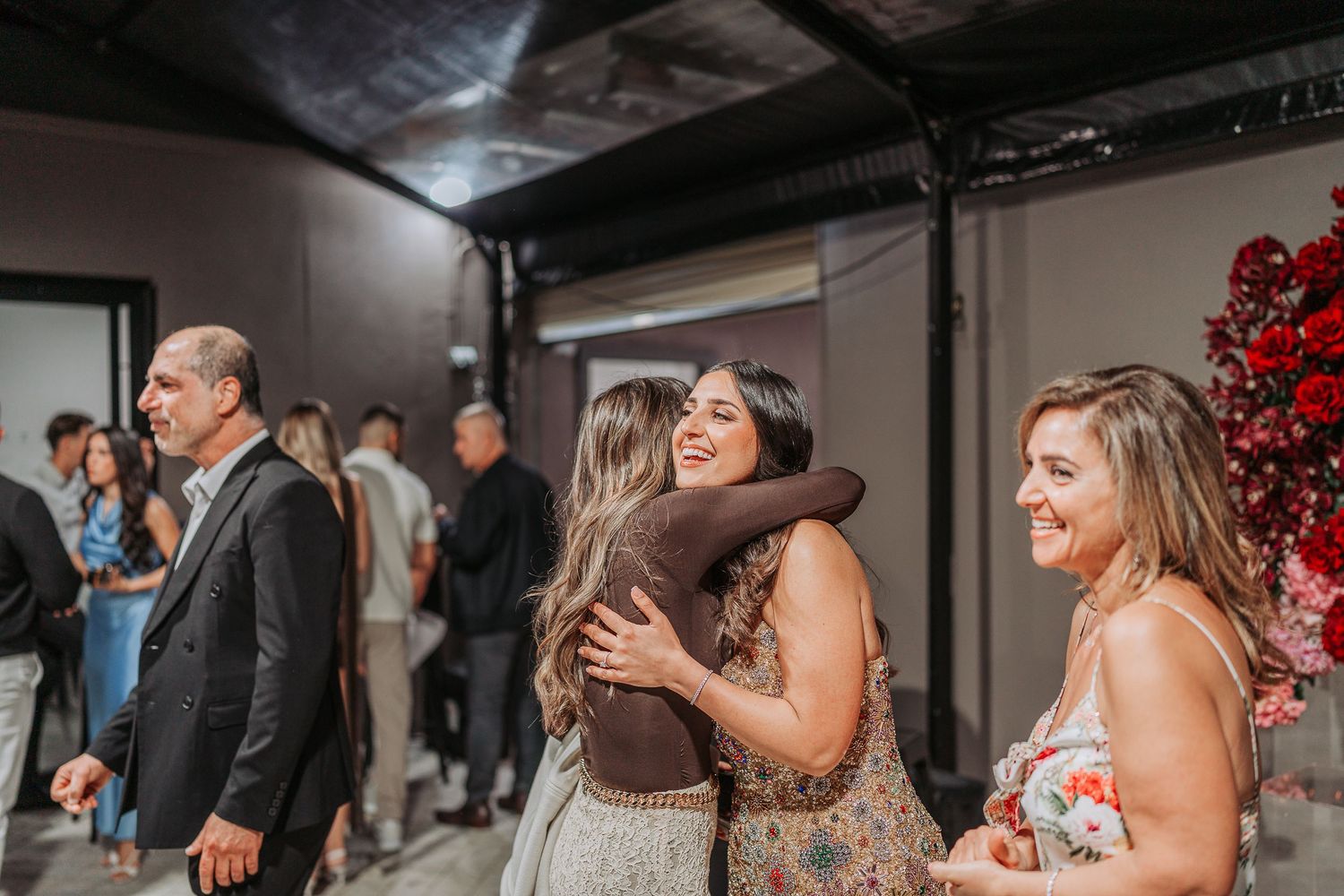 Joyful celebration moment with guests hugging near red floral decorations.