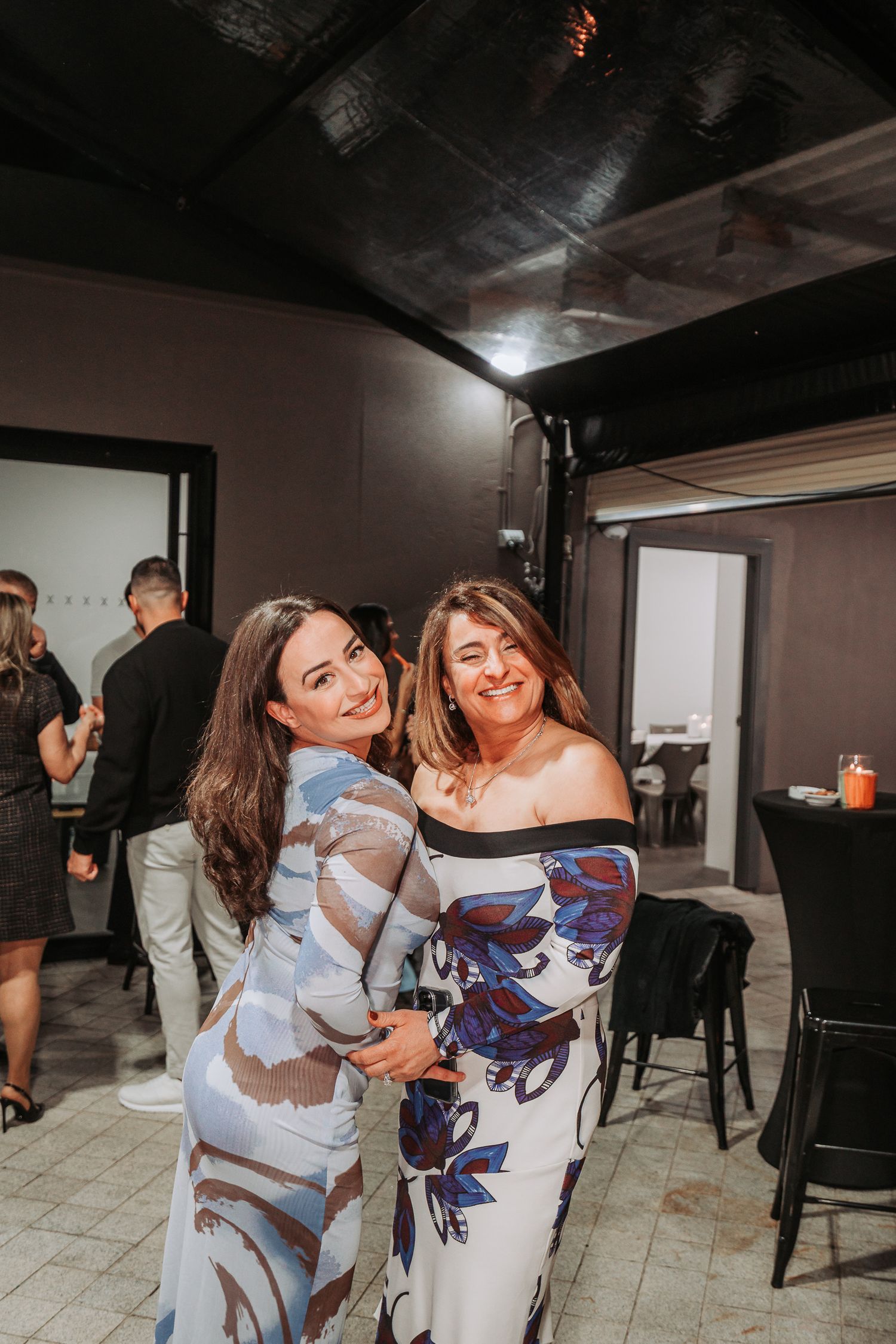 Two women in floral dresses embrace and smile at a celebration event.