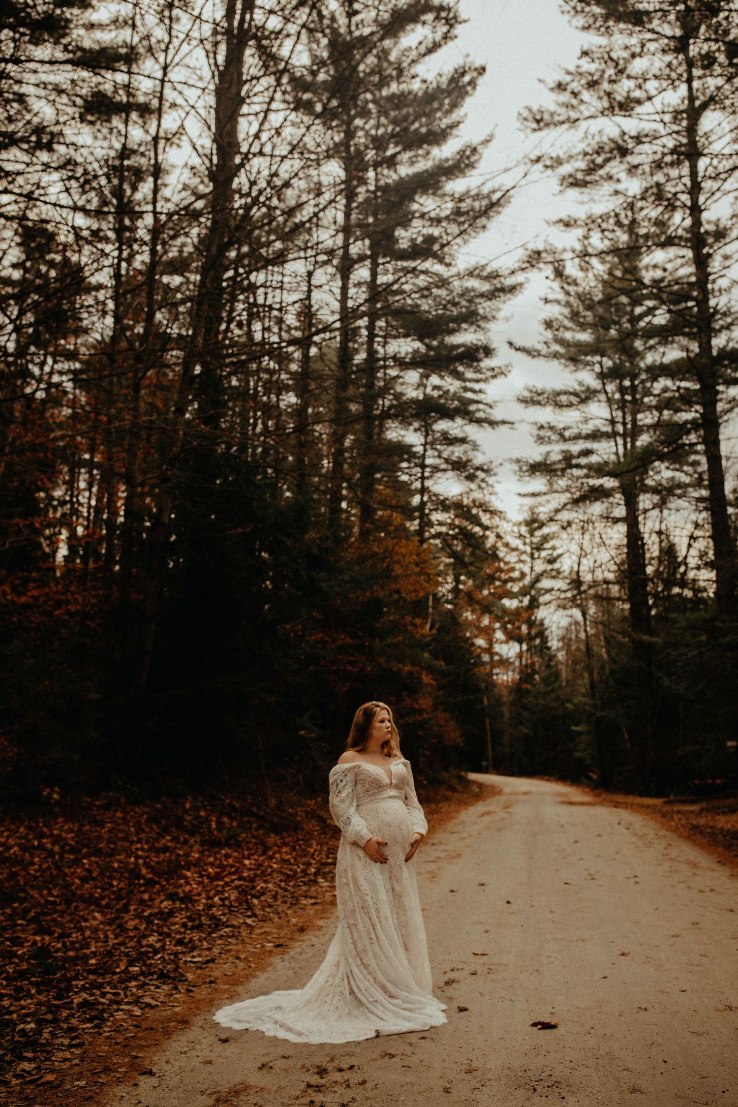 A flowing white dress catches the light on a forest path covered in autumn leaves.