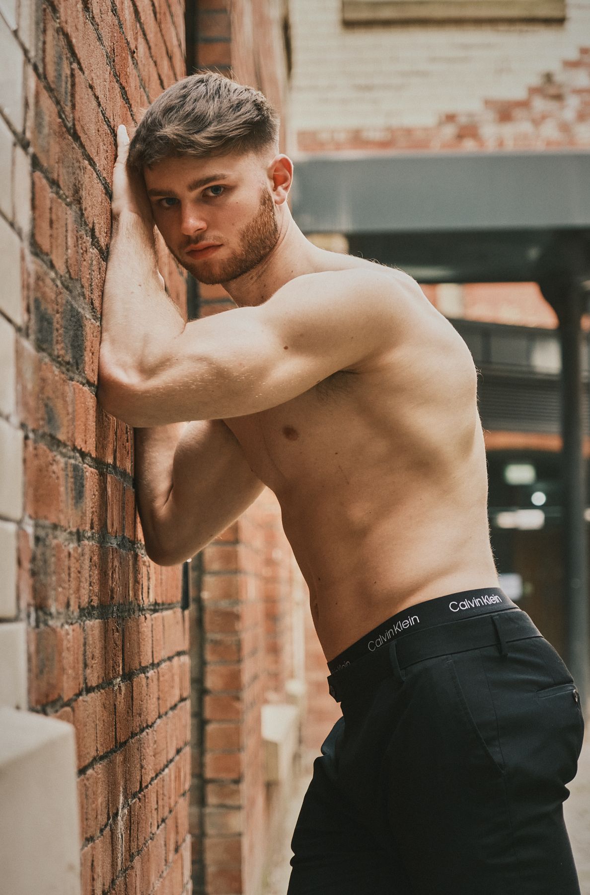 Shirtless male model leaning against a brick wall, gazing into the camera with a serious expression