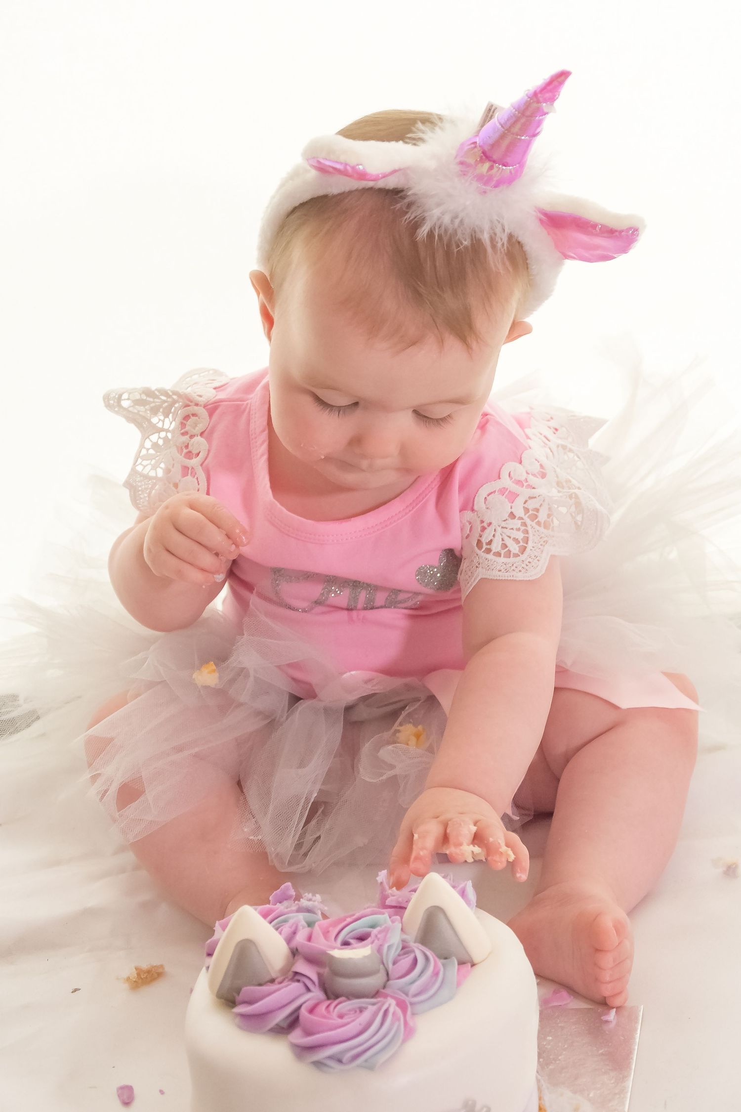 Baby in pink tutu and unicorn headband reaching for cake decorations.