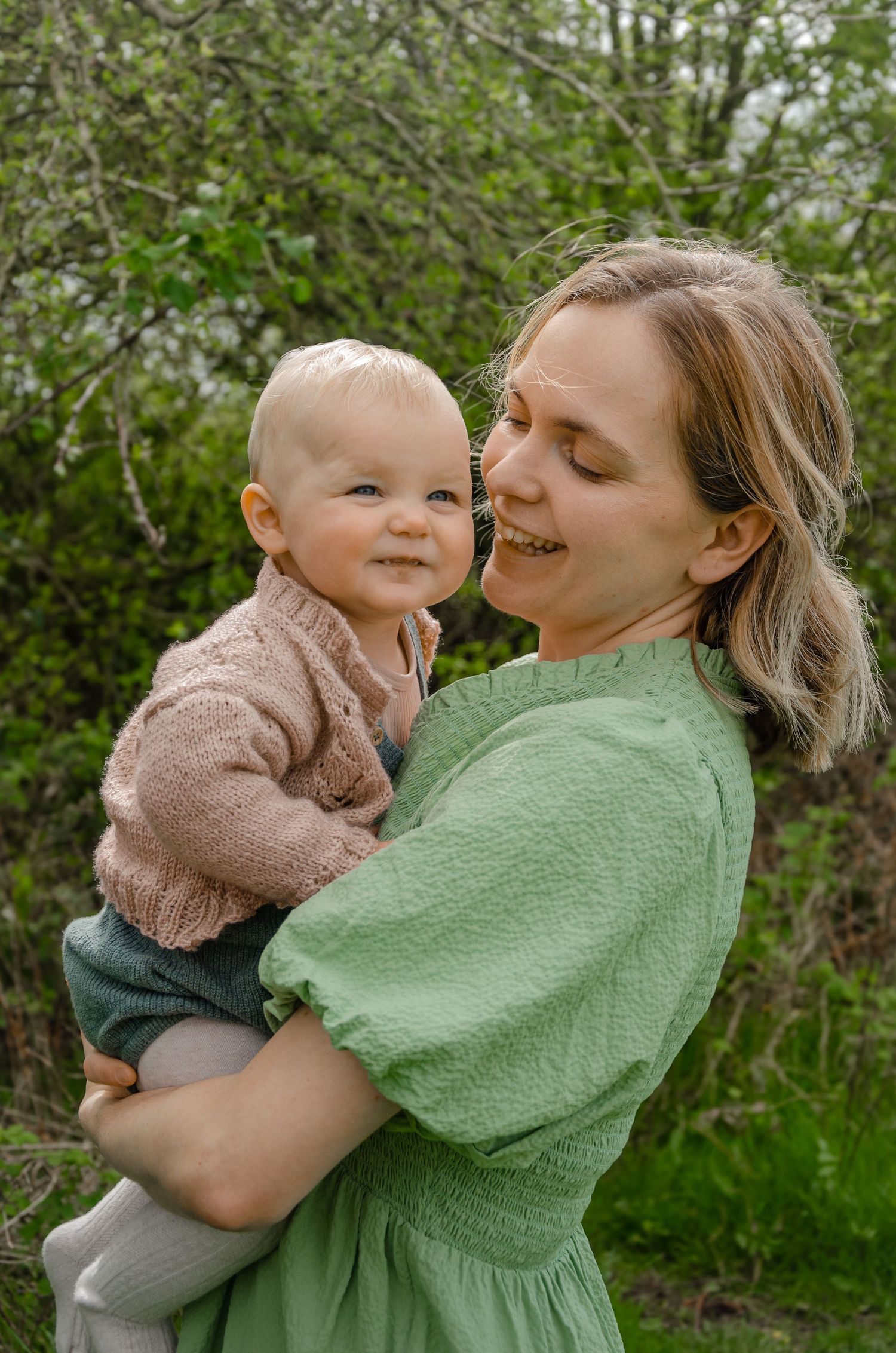 Parent and baby share a tender moment outdoors in springtime greenery.
