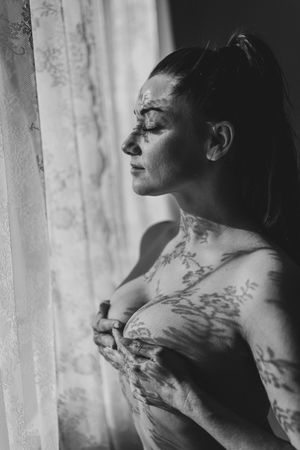 A moody black and white artistic portrait captures a figure with tattoos bathed in soft window light.
