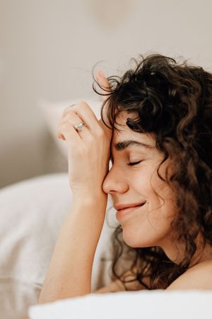 Someone with dark curly hair lies peacefully on white bedding with a gentle smile and closed eyes, suggesting relaxation.