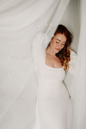 Elegant figure in a fitted white dress poses gracefully against a white backdrop with eyes closed and flowing auburn hair.