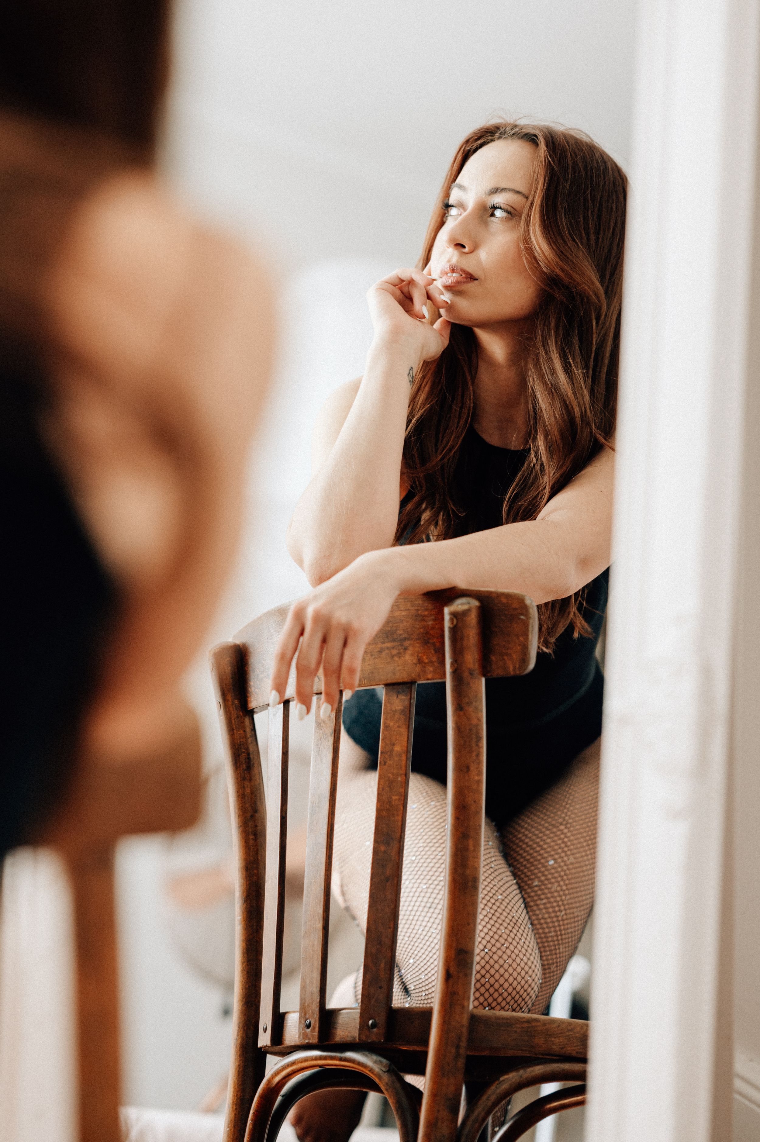 A woman in a black outfit poses thoughtfully on a wooden chair in a warm-toned artistic portrait series.