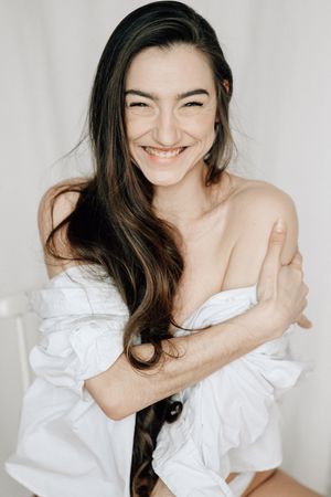 Woman in white draped fabric laughs joyfully while posing against white background in portrait series.