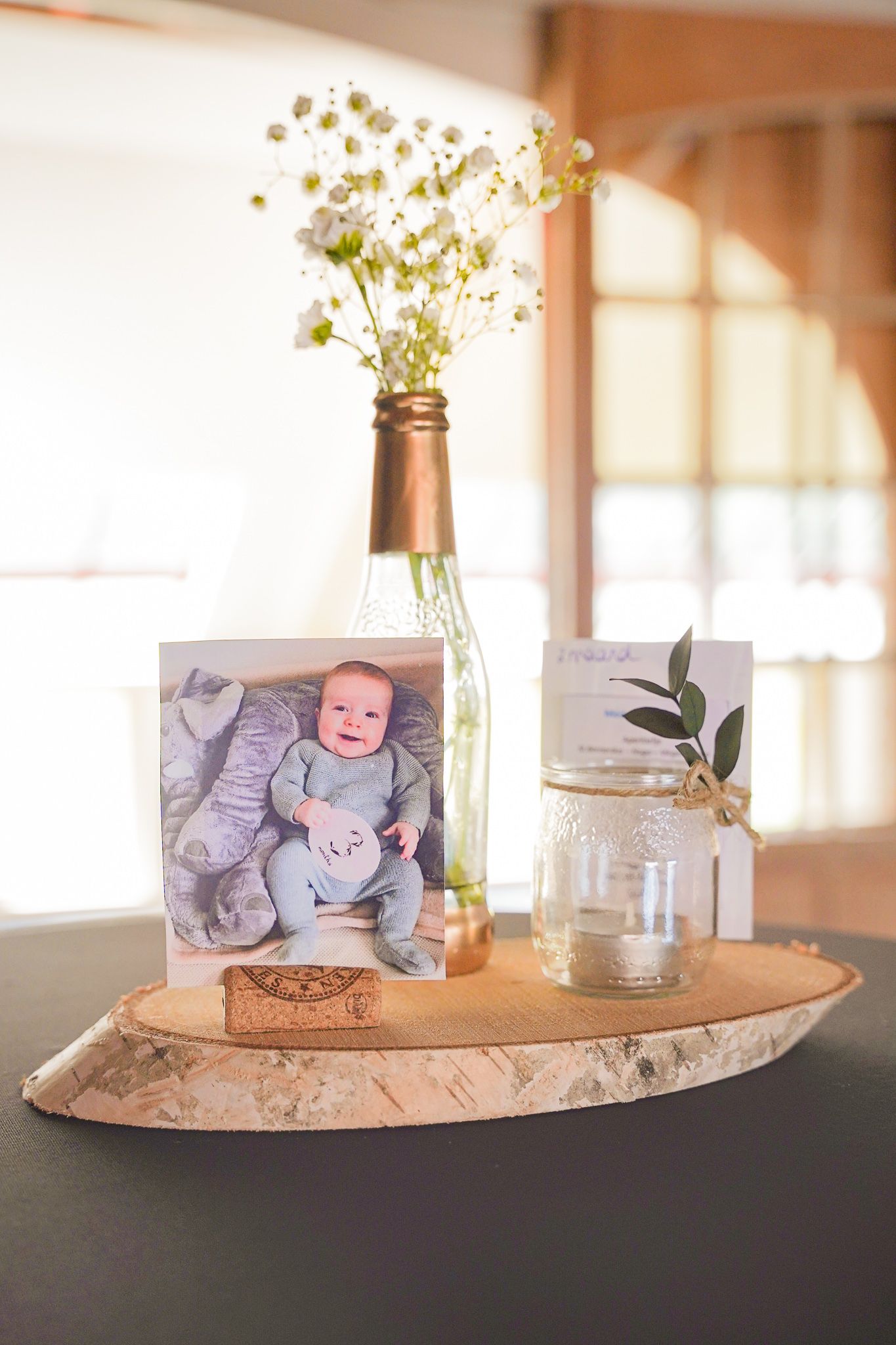 Simple centerpiece with baby photo, wildflowers in bottle, and jar on wooden slice.